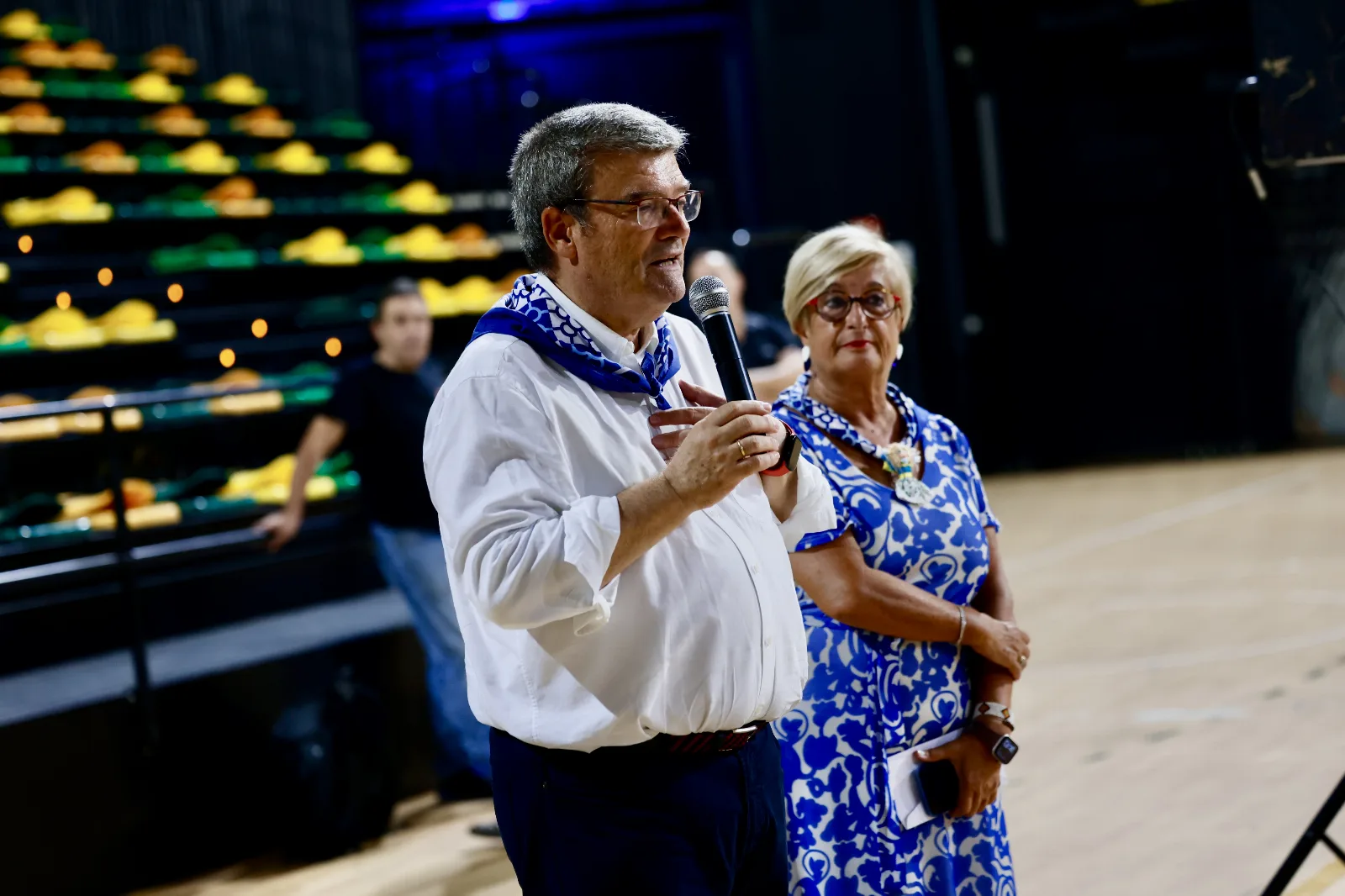 El Alcalde de Bilbao, con pañuelo azul al cuello, habla por un micrófono de mano en la pista del pabellón acompañado por otra persona