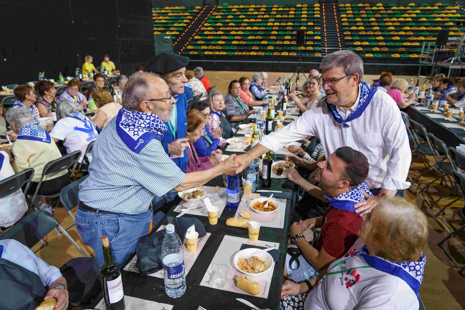 Un comensal se levanta para estrechar la mano por encima de la mesa larga, con los platos servidos, el pan, el agua y las botellas de vino delante