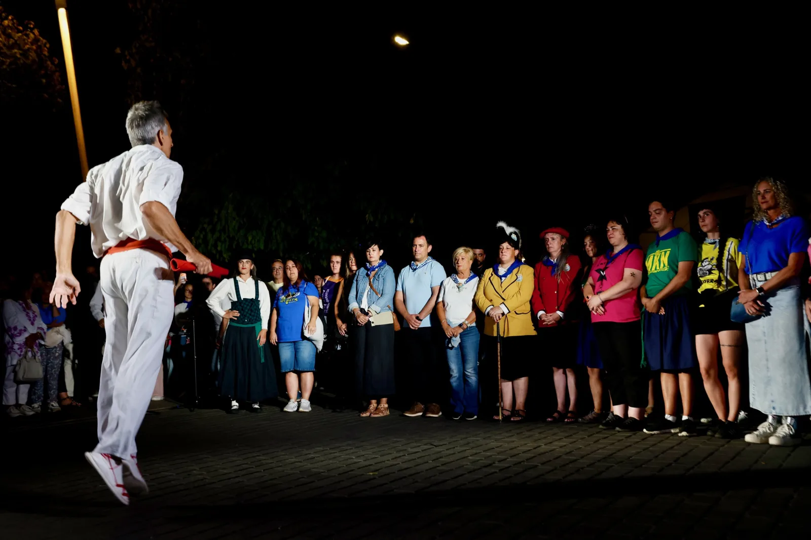 Un dantzari vestido de blanco con faja roja salta en el aire durante un aurresku, de noche, ante una fila de personas que lo observan