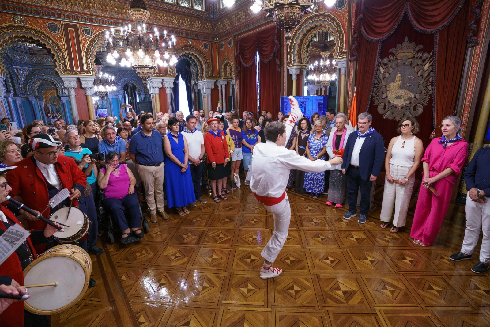 Un dantzari vestido de blanco con faja roja baila en el centro del Salón Árabe, rodeado por los asistentes en pie