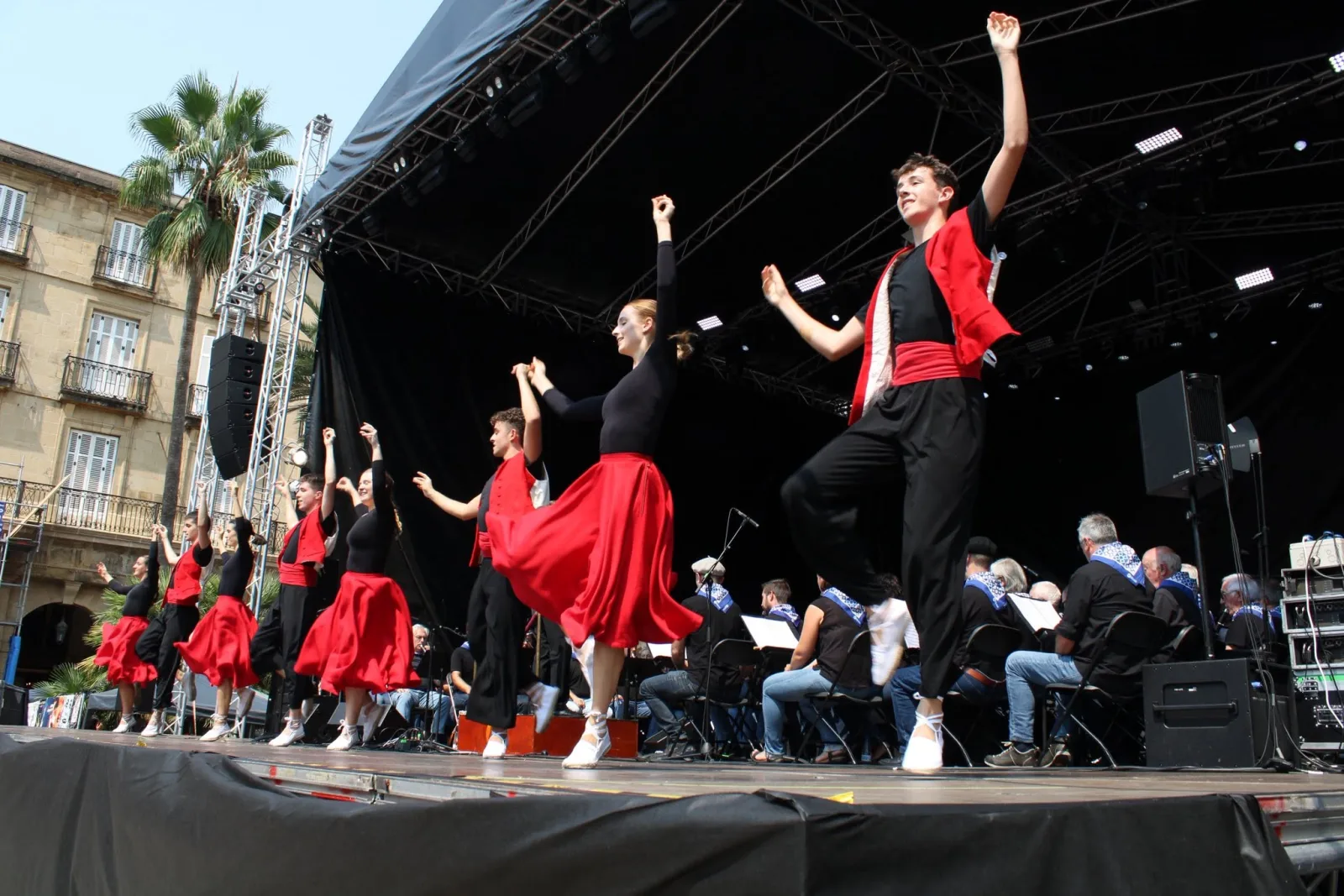Un grupo de dantzaris de negro y rojo salta con los brazos en alto sobre el escenario, con la banda sentada detrás
