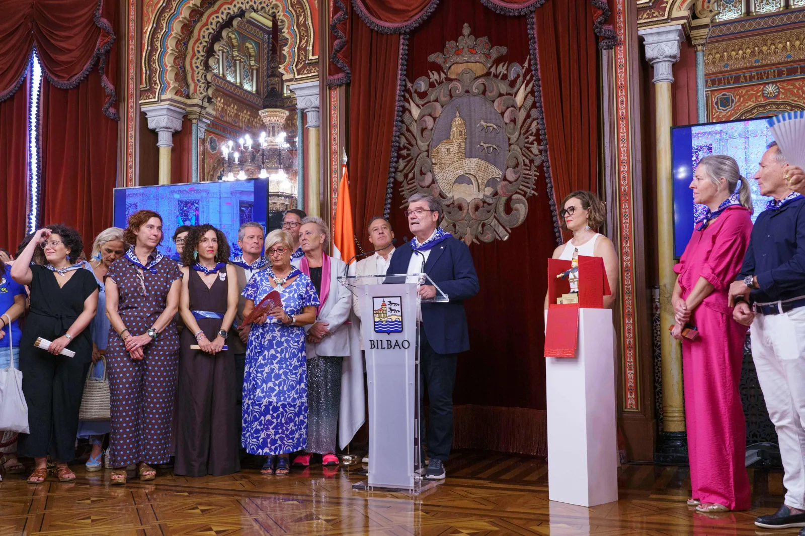 El Alcalde de Bilbao habla desde un atril con el escudo de la villa mientras un grupo de asistentes escucha a los lados