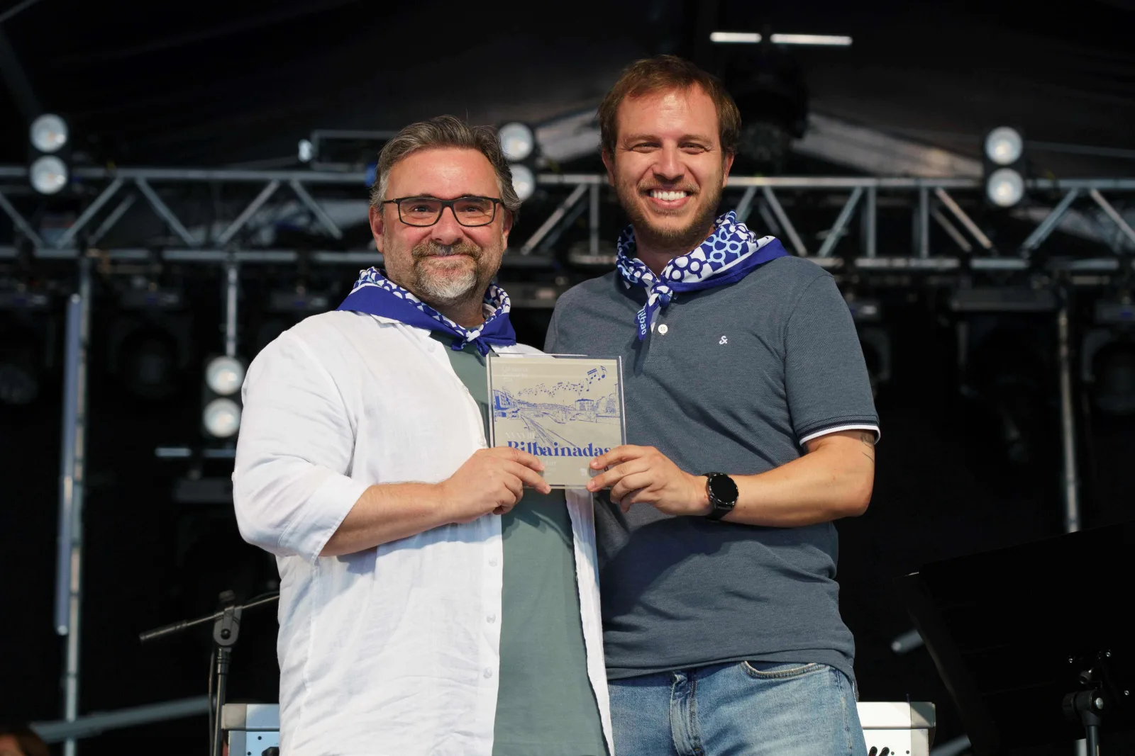 Dos hombres con pañuelo azul sostienen juntos la placa del concurso mirando a cámara