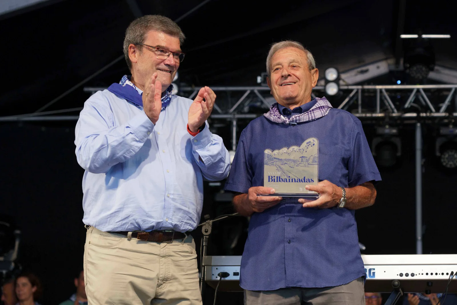 El Alcalde de Bilbao, con camisa celeste y pañuelo azul, aplaude junto a un hombre que sostiene una placa de metacrilato con la palabra Bilbainadas