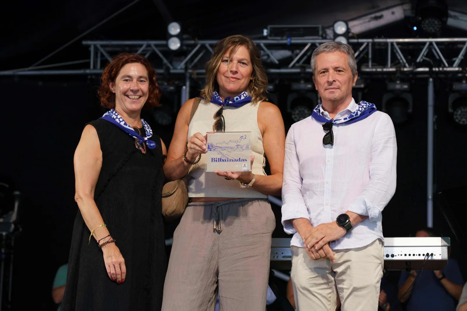 Tres personas con pañuelo azul posan de pie; la del centro sostiene con las dos manos la placa del concurso
