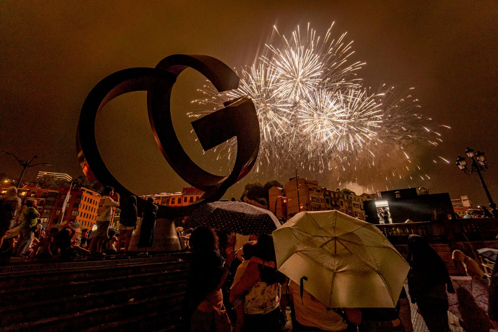 Una gran escultura circular de acero ocupa la izquierda de la imagen mientras a su derecha estallan varios fuegos artificiales blancos; abajo, el público bajo paraguas