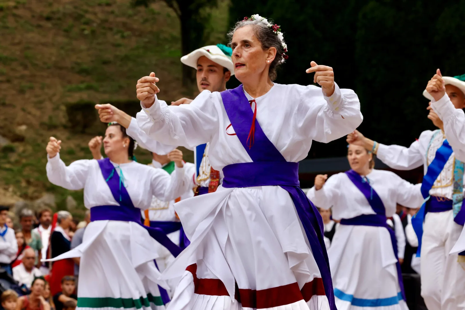 Varias dantzaris con blusa blanca, falda blanca y banda morada bailan con los brazos levantados