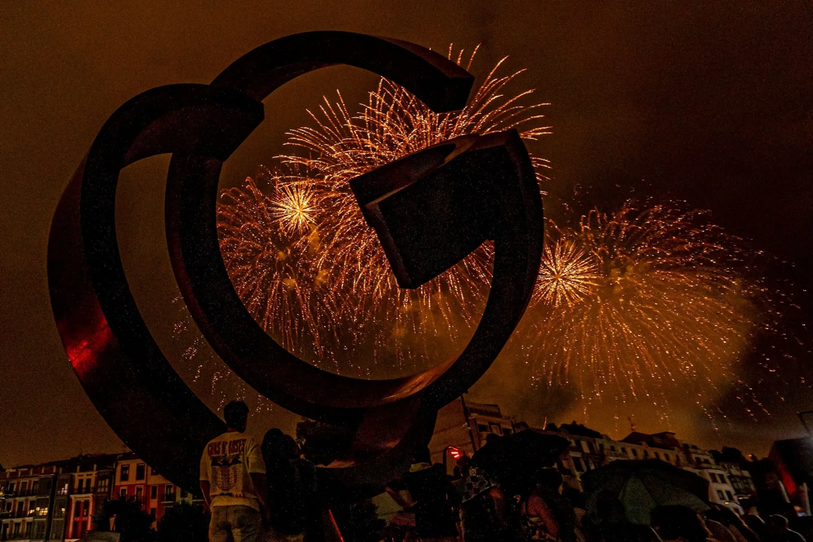 Fuegos artificiales dorados vistos a través del hueco de una gran escultura circular de acero, con público en la parte baja de la imagen