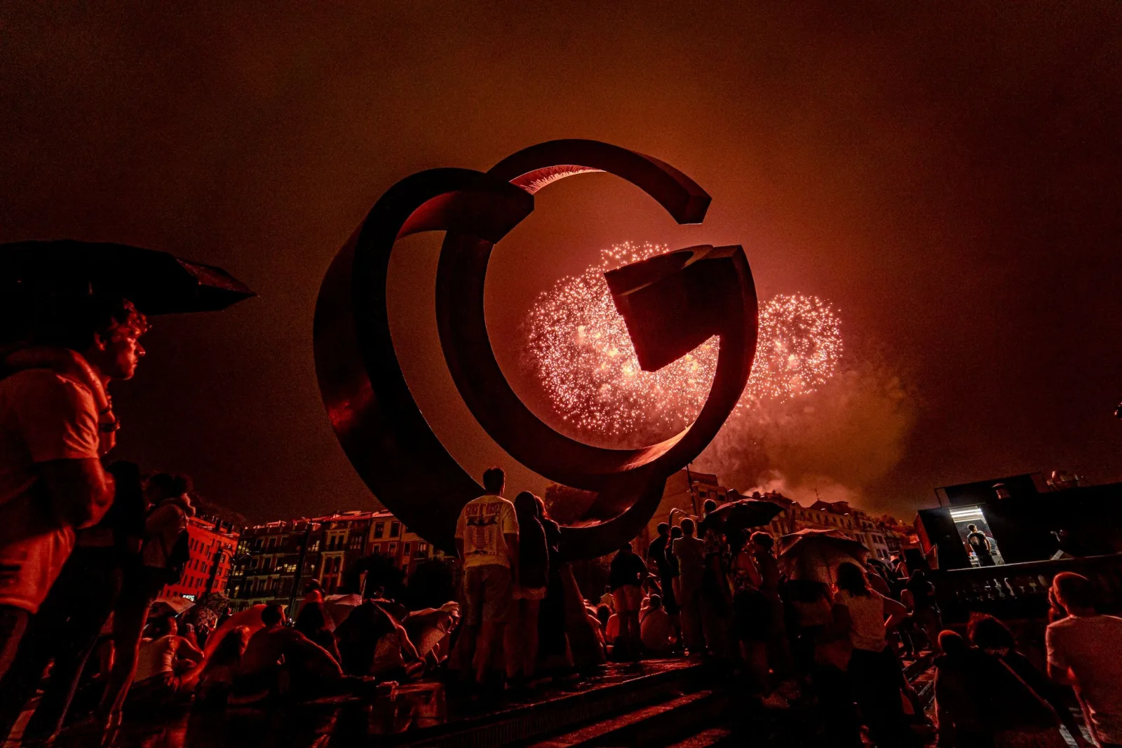 Un estallido rosado de fuegos artificiales se ve dentro del hueco de una escultura circular de acero mientras el público lo mira desde las escaleras del muelle
