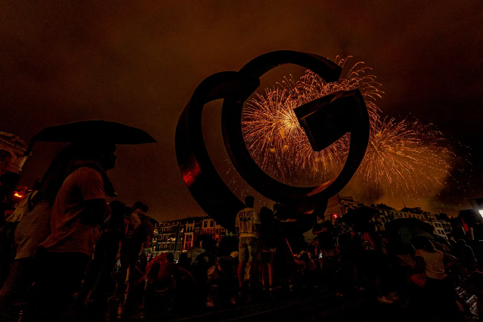 La silueta oscura de una gran escultura de acero en primer plano, con dos abanicos de chispas doradas estallando justo detrás