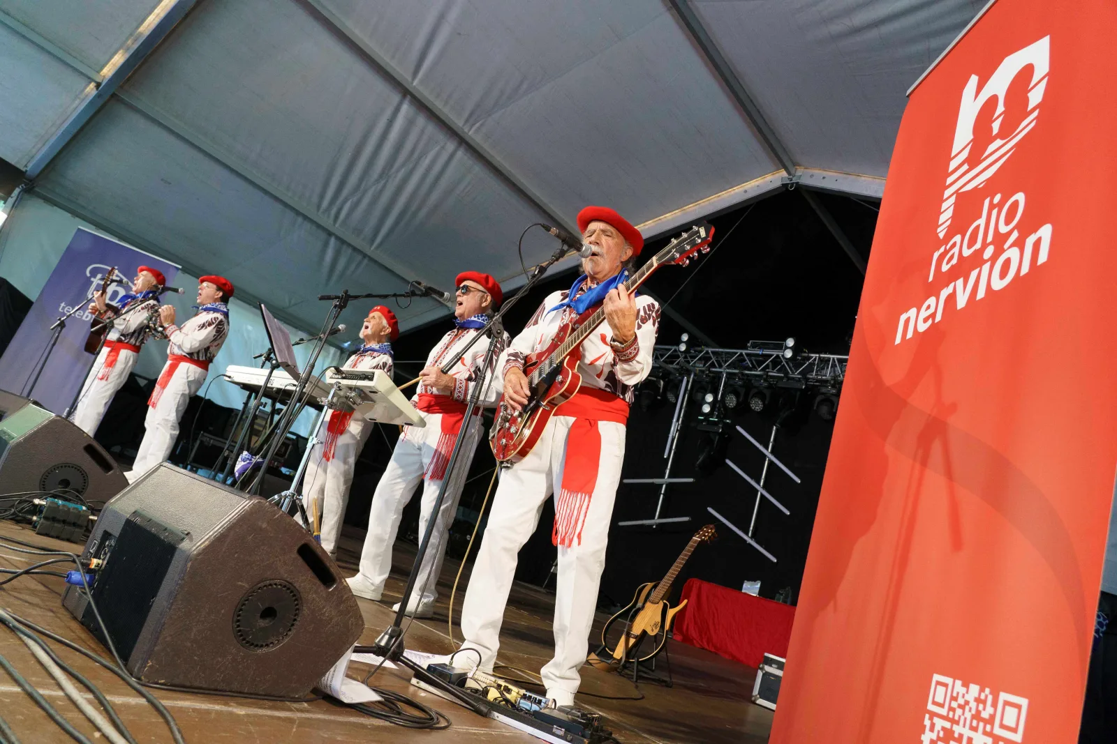 Seis músicos con traje blanco de detalles rojos y txapela roja tocan sobre el escenario cubierto de la Pérgola, junto a una lona vertical de Radio Nervión