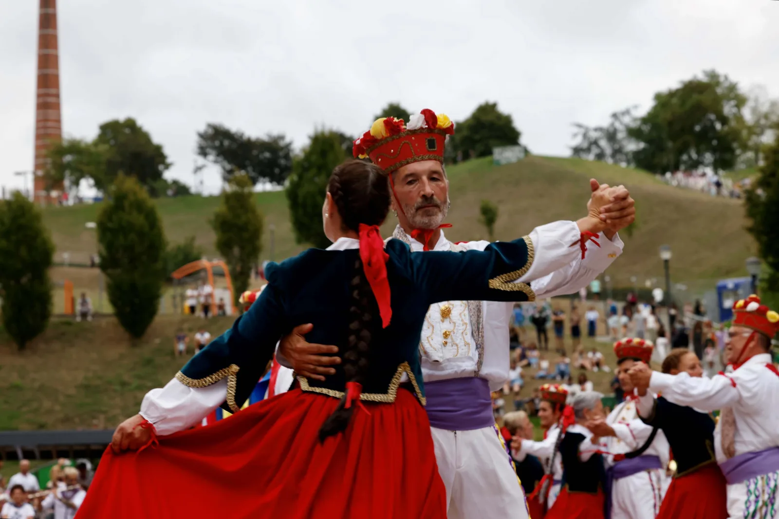 Una pareja con traje tradicional baila agarrada, con la chimenea de ladrillo del parque al fondo