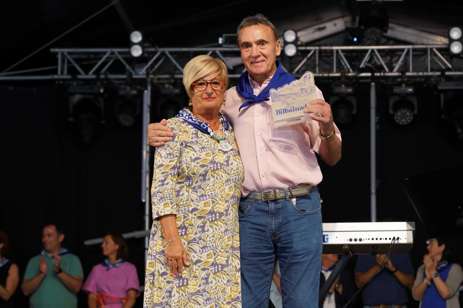 Una mujer con vestido estampado y un hombre con camisa rosa y pañuelo azul posan abrazados mientras él sostiene en alto la placa del concurso