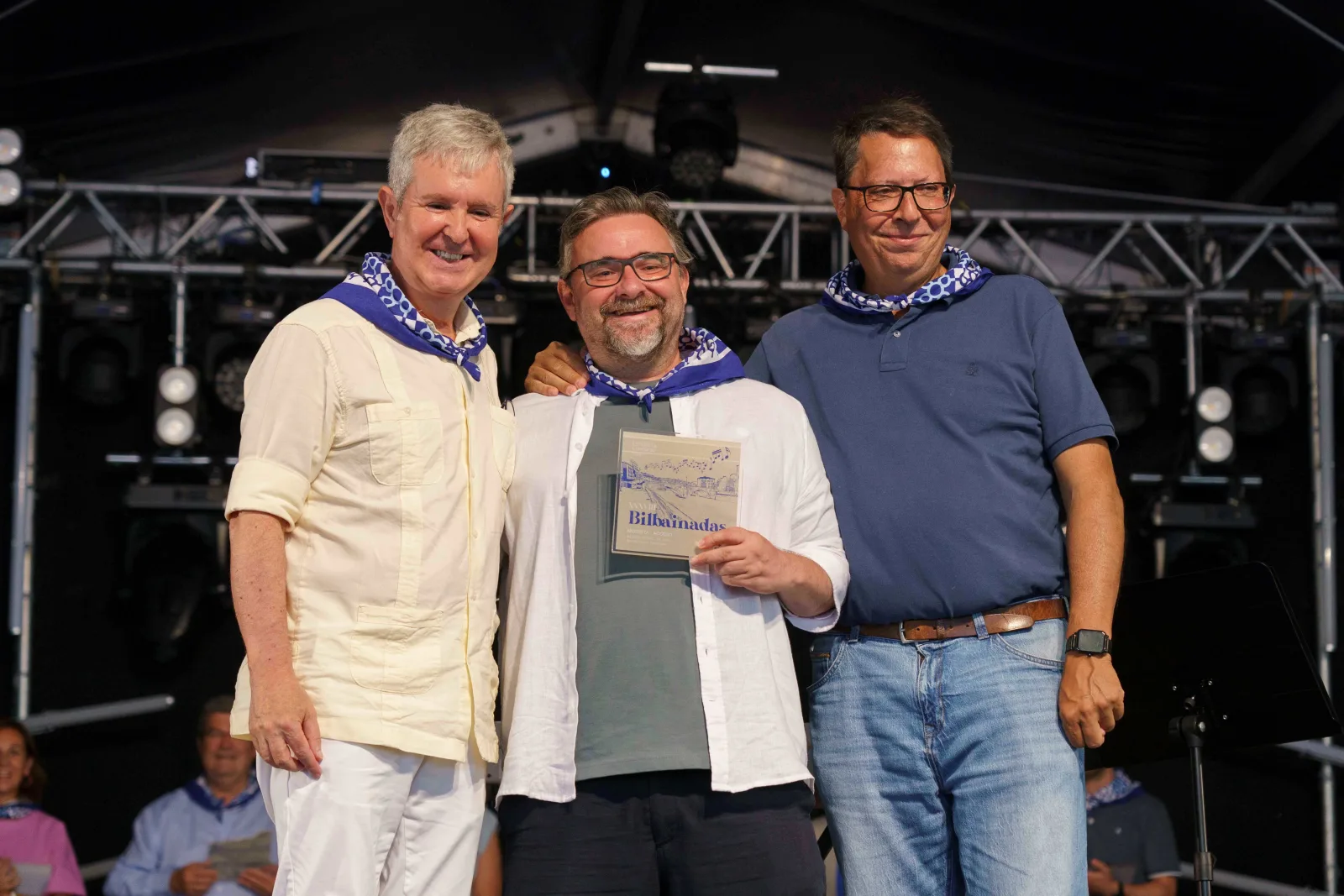 Tres hombres posan abrazados sobre el escenario; el del centro sostiene la placa del concurso
