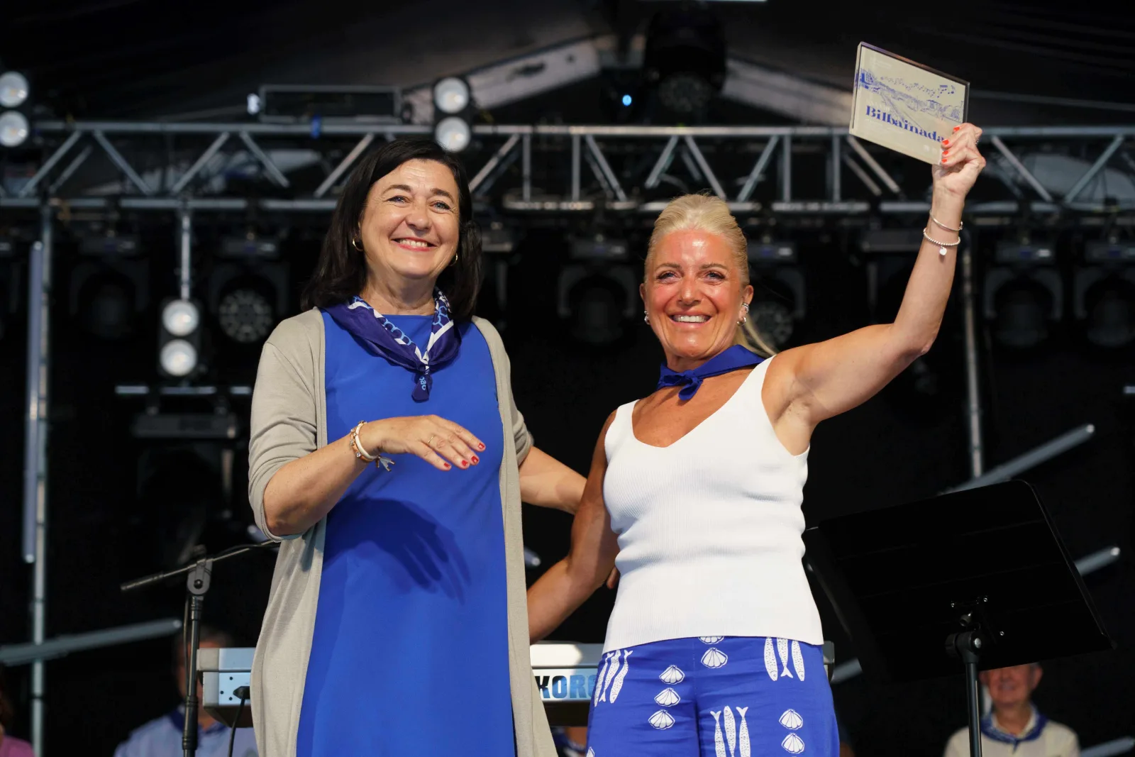 Dos mujeres posan sobre el escenario; la de la derecha levanta con el brazo estirado la placa del concurso