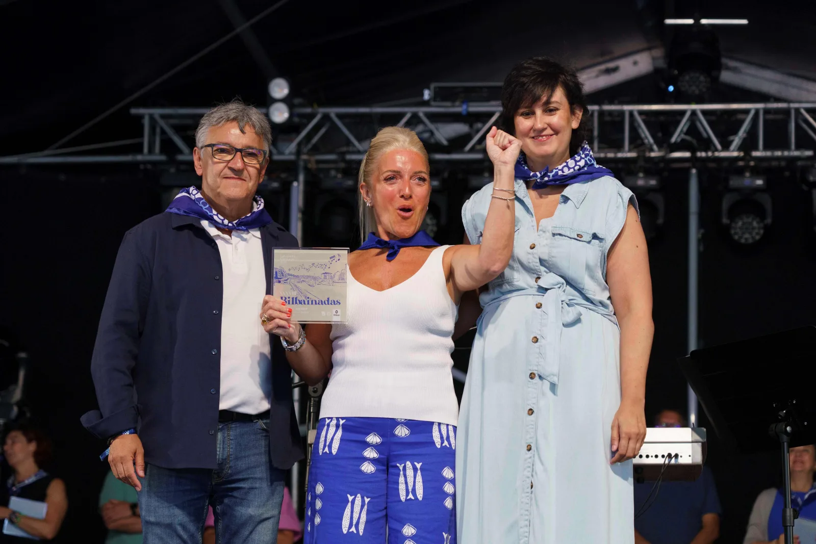 Tres personas posan sobre el escenario; la del centro sostiene la placa del concurso y levanta el puño
