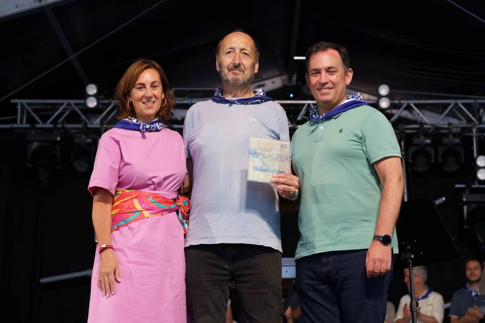 Tres personas posan de pie con pañuelo azul; el del centro sostiene la placa del concurso a la altura del pecho