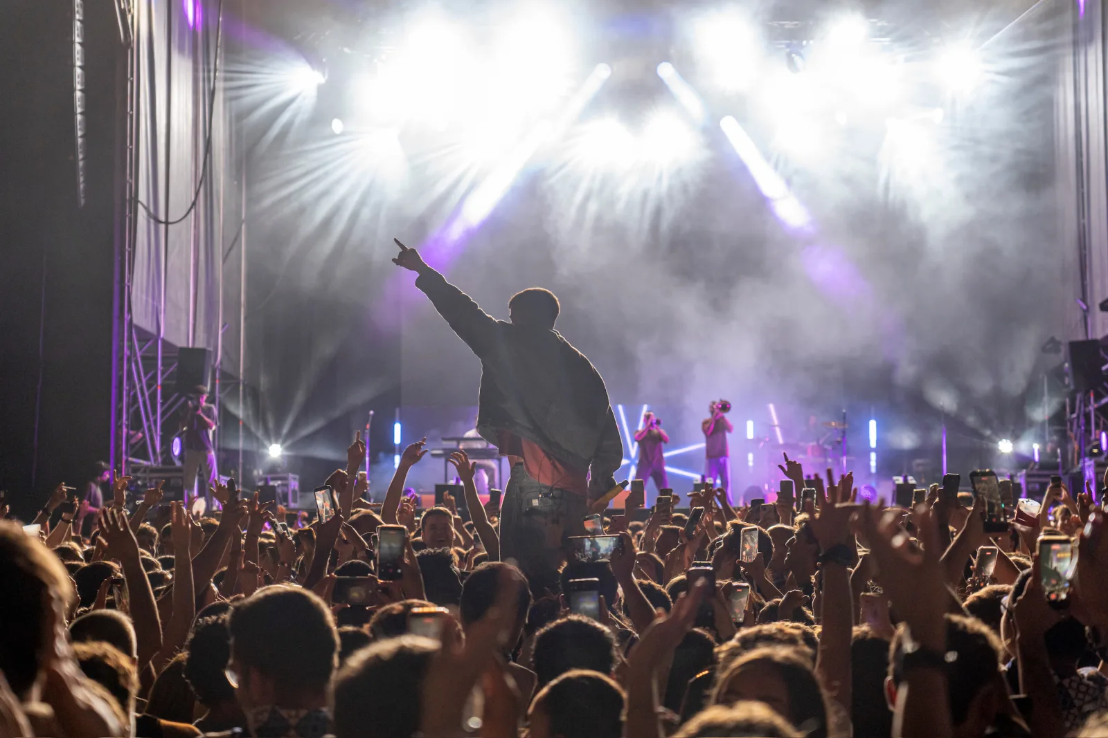 Una persona a hombros entre el público señala hacia el escenario, rodeada de móviles en alto y focos blancos