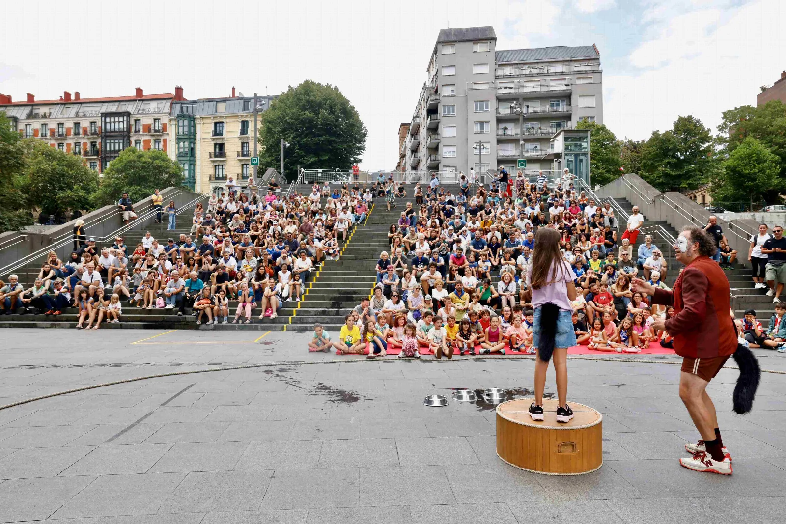 Cientos de personas sentadas en unas escaleras de hormigón ven un espectáculo de calle en el que dos artistas actúan sobre una tarima circular