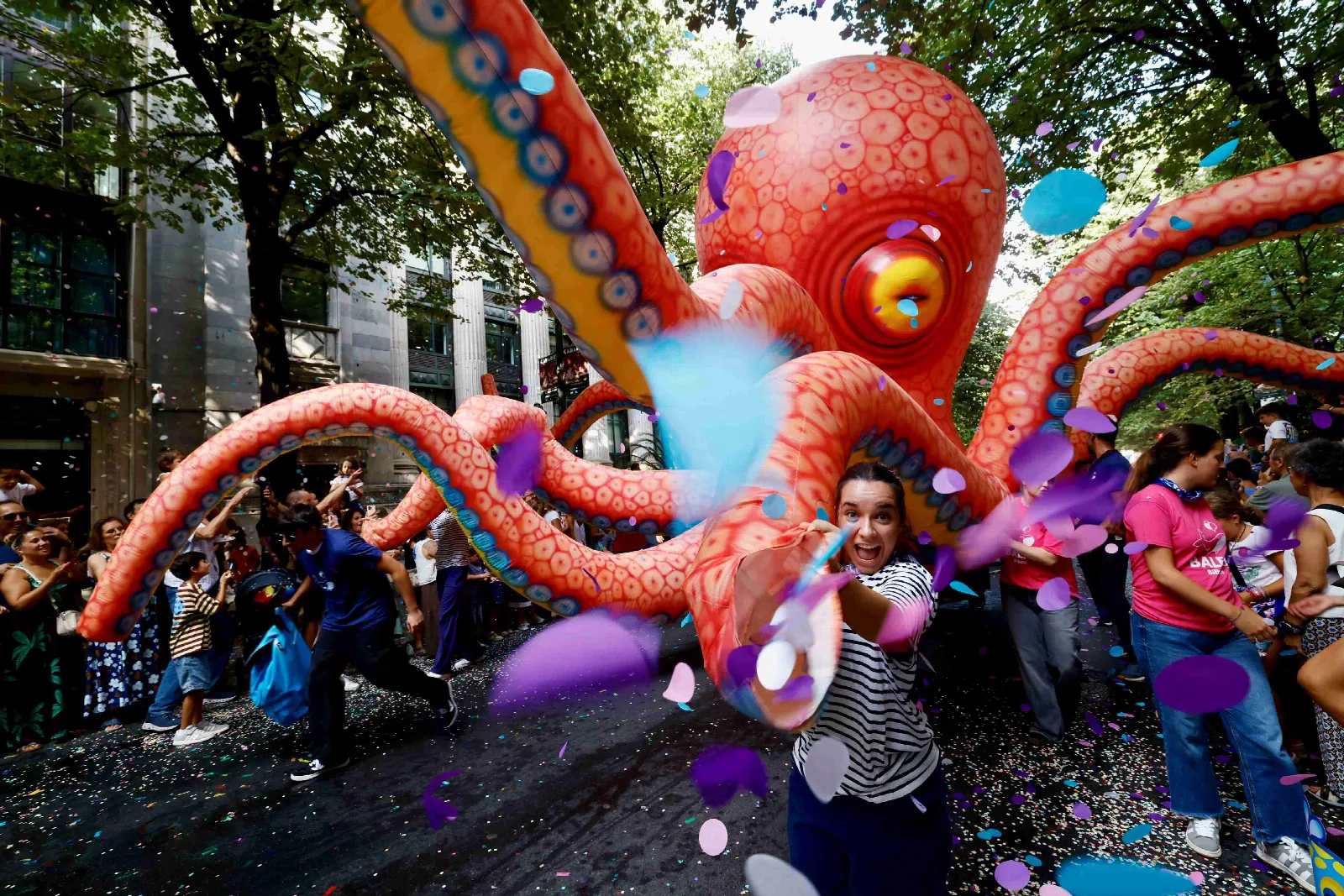 Un pulpo gigante hinchable de color naranja avanza por una calle arbolada de Bilbao entre confeti y globos, rodeado de público
