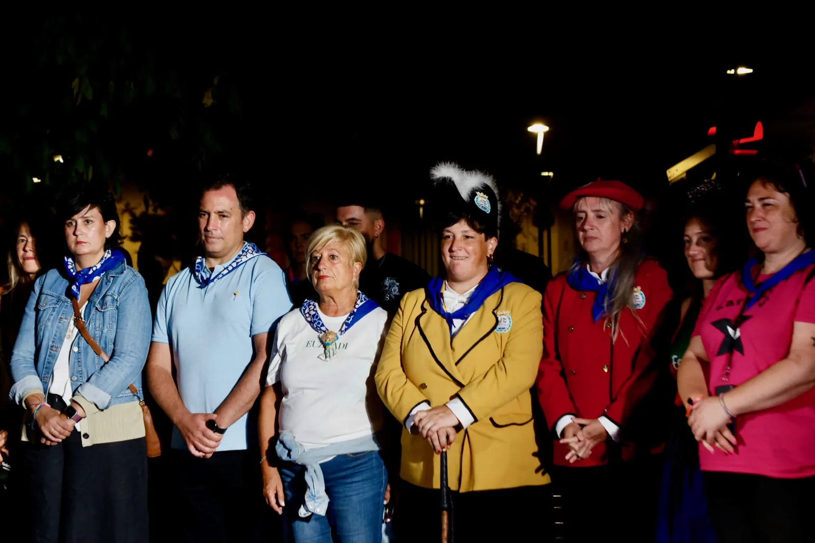 Fila de personas con pañuelos azules al cuello y camisetas de comparsa observan un acto de noche; entre ellas, dos mujeres con las chaquetas de gala amarilla y roja del Ayuntamiento de Bilbao