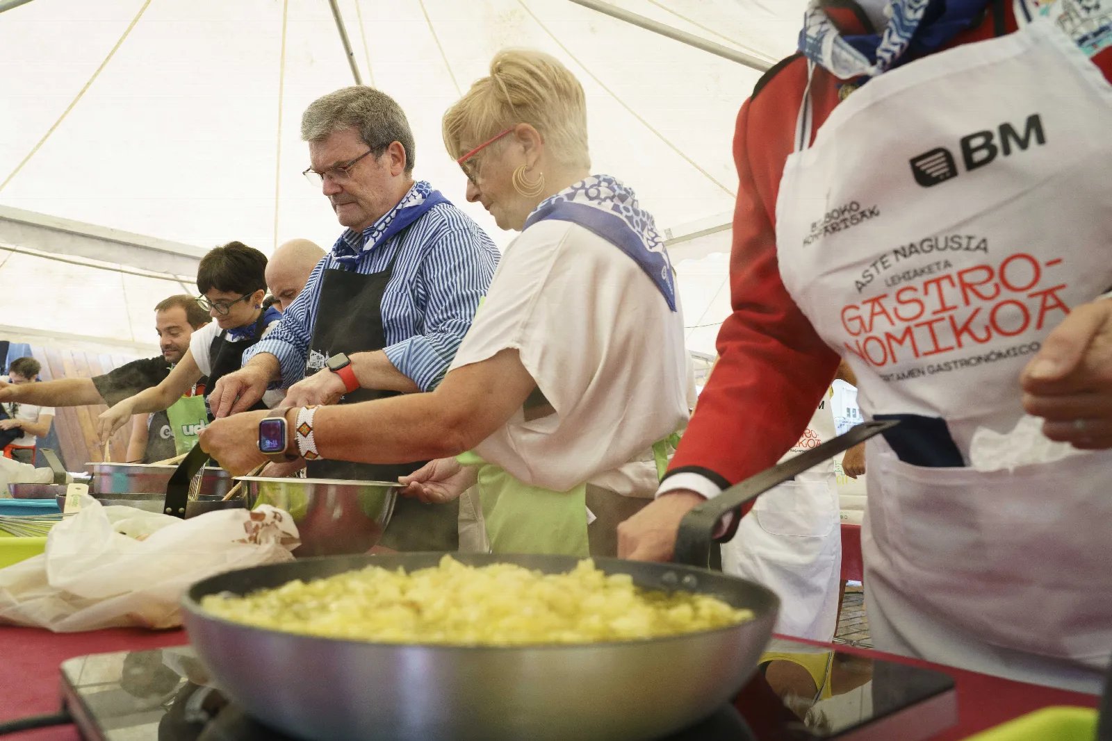Sartén en primer plano con patata cortada haciéndose, y participantes trabajando al fondo