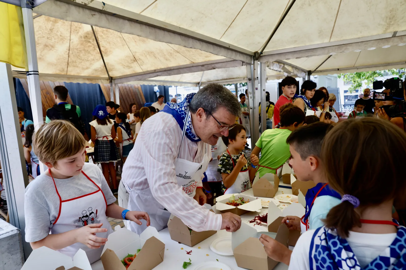 Un cocinero con delantal y pañuelo atiende a varios niños alrededor de una mesa del taller, bajo la carpa del Arenal