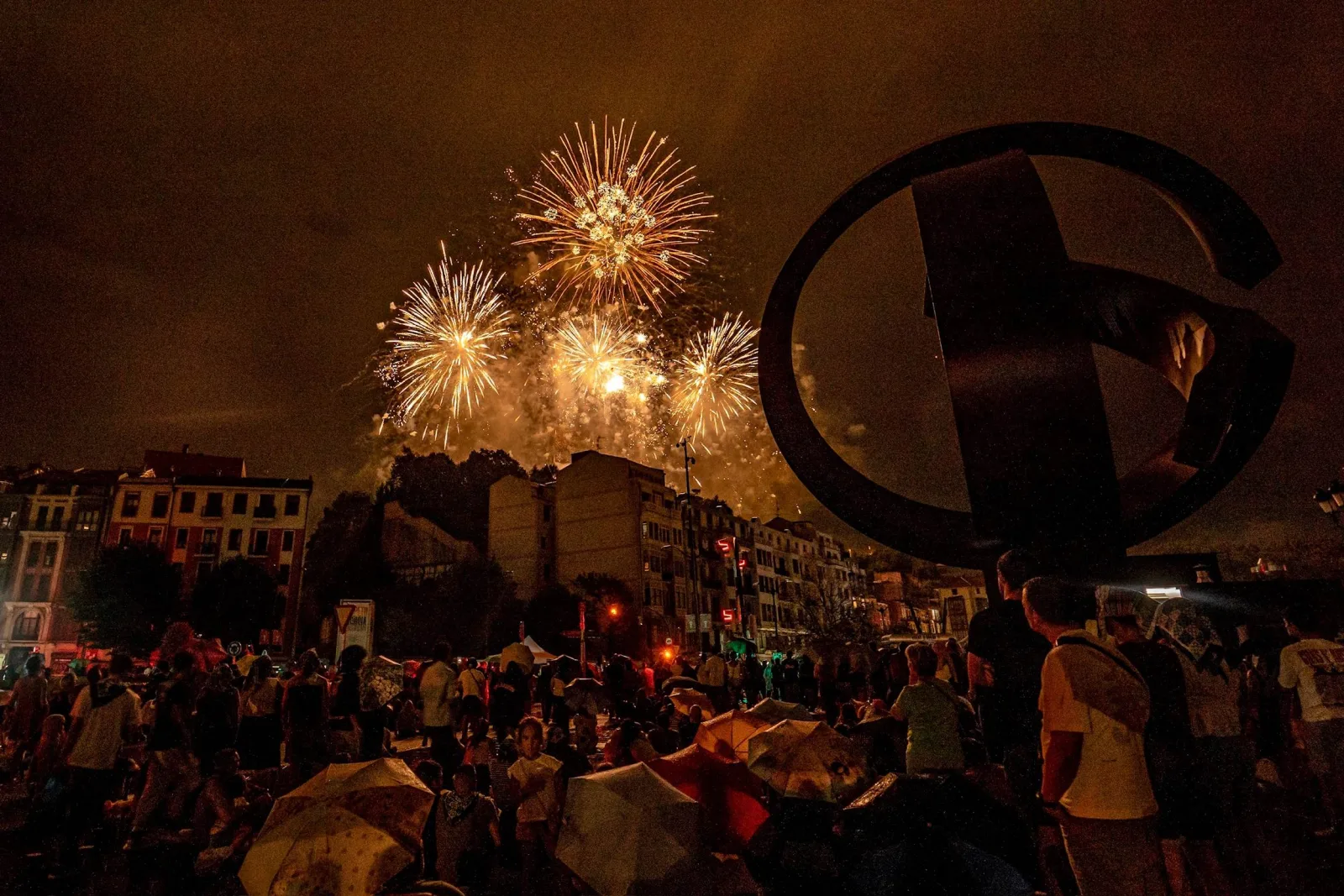 Varias palmeras doradas de fuegos artificiales estallan sobre los edificios de la ría de Bilbao, con una gran escultura de acero en primer plano a la derecha y el público con paraguas abajo