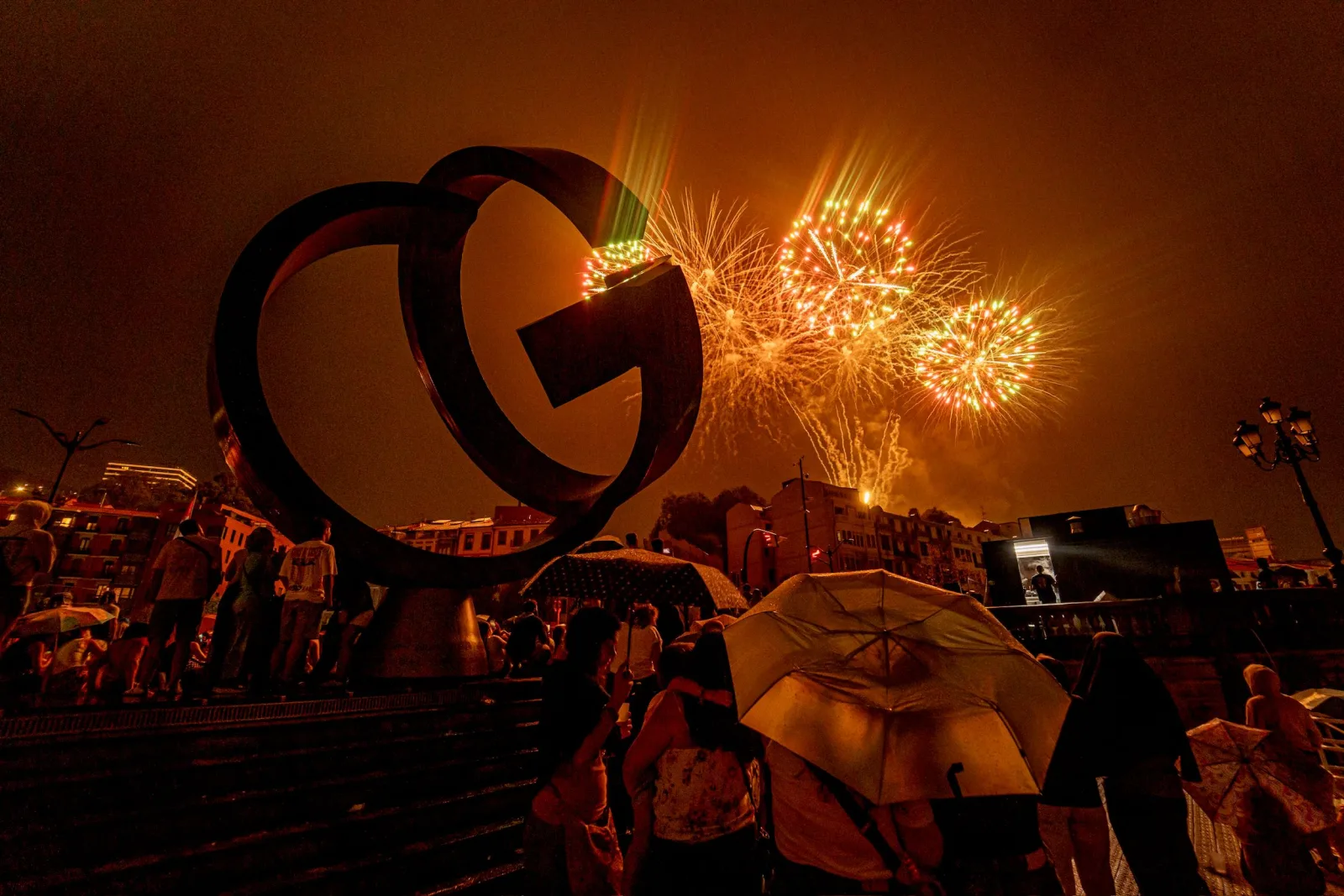 Fuegos artificiales verdes y dorados estallan a la derecha de una gran escultura de acero mientras el público mira desde abajo bajo paraguas