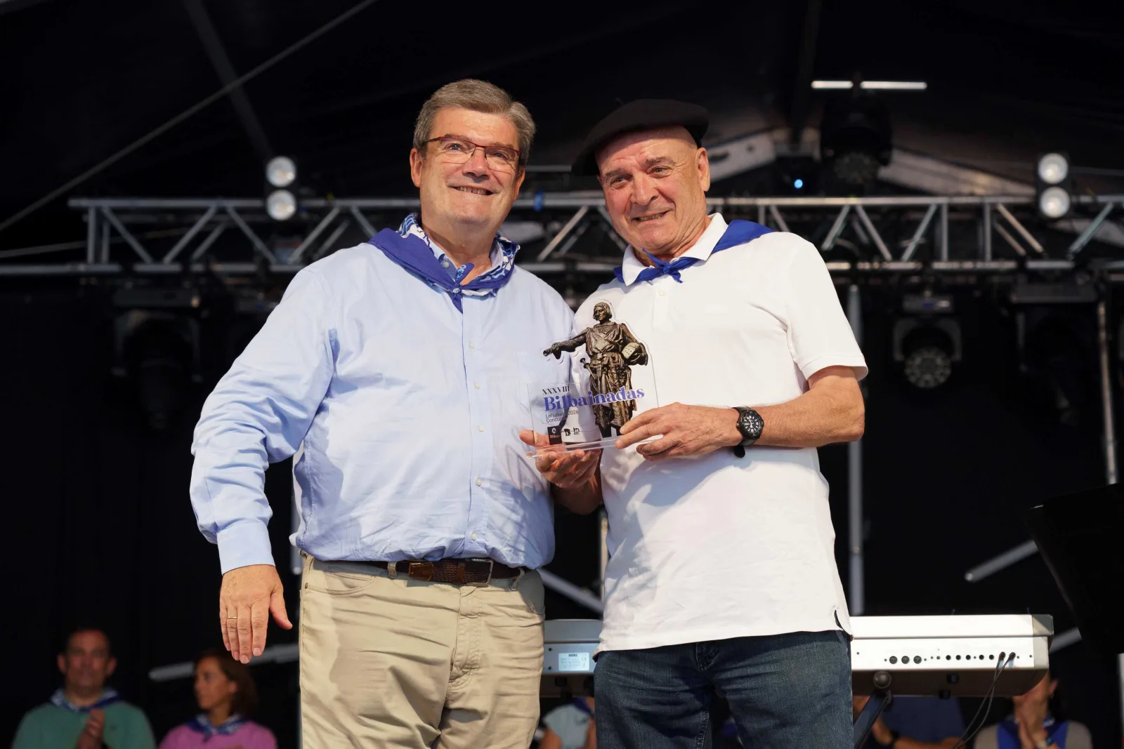 Dos hombres posan sobre el escenario sosteniendo juntos un trofeo formado por una figura sobre peana y una placa con la palabra Bilbainadas; el de la derecha lleva txapela negra