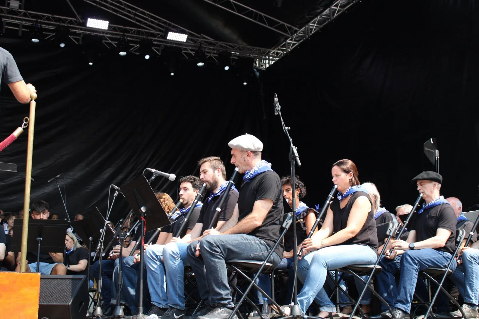 Una fila de txistularis sentados en el escenario toca el txistu con pañuelo azul al cuello, con los atriles delante