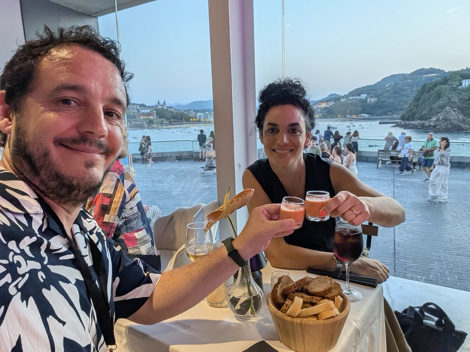 Esteban y Elena brindando con los vasos de salmorejo en su mesa junto al ventanal del Aquarium, con la bahía y el Paseo Nuevo llenos de gente al otro lado del cristal