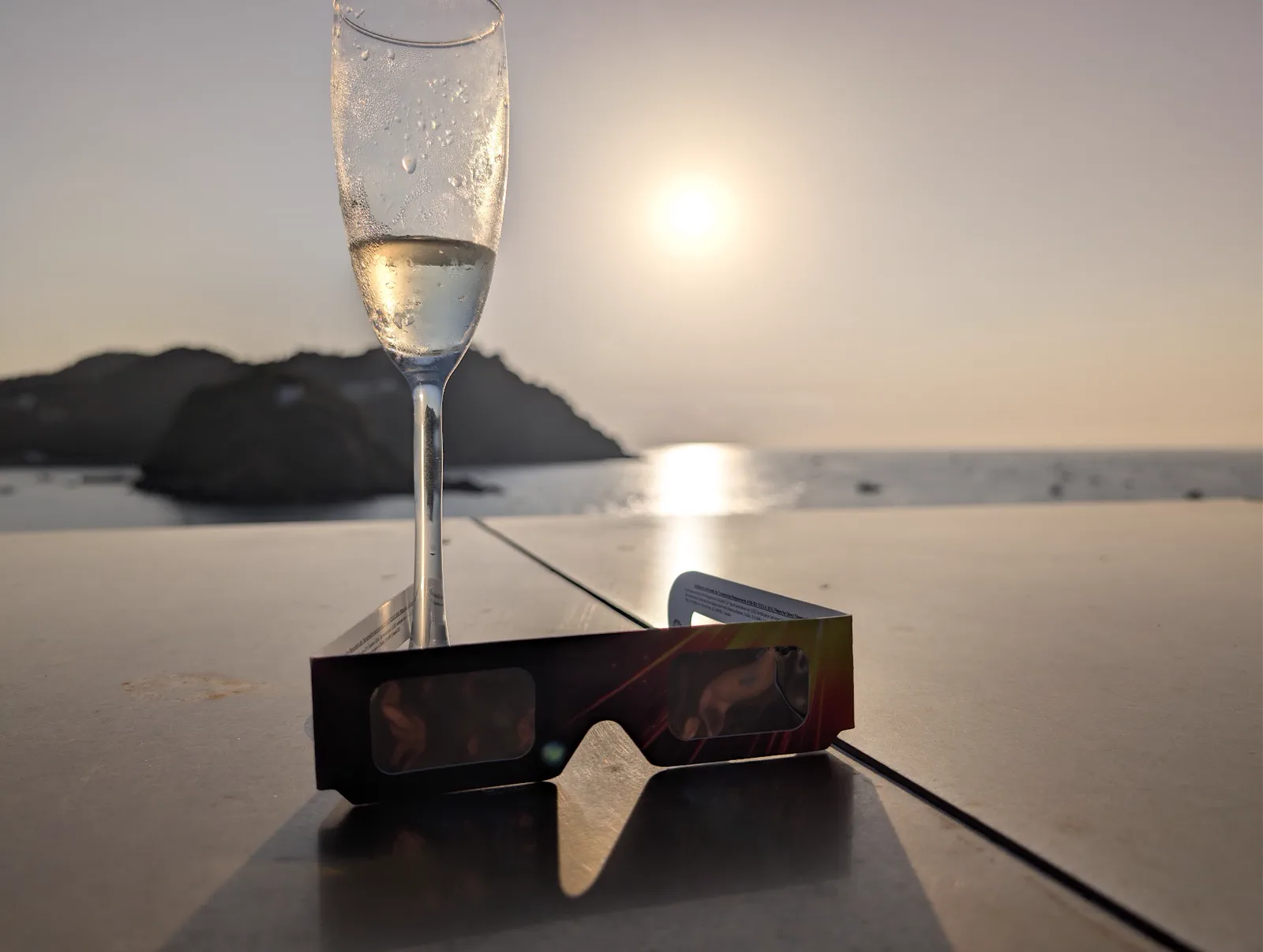 Una copa de cava y unas gafas de eclipse apoyadas en el muro de la terraza del Aquarium, con la isla de Santa Clara y el sol bajo sobre el mar al fondo