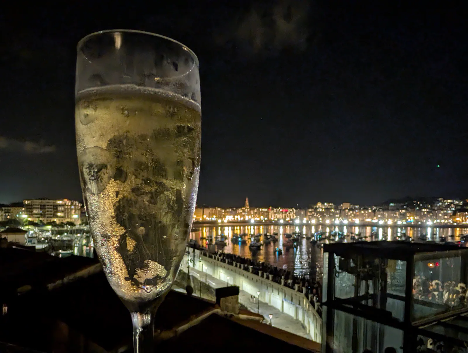 Copa de champán en primer plano y, al fondo, la bahía de la Concha ya de noche con la ciudad iluminada, los barcos fondeados y el Paseo Nuevo lleno