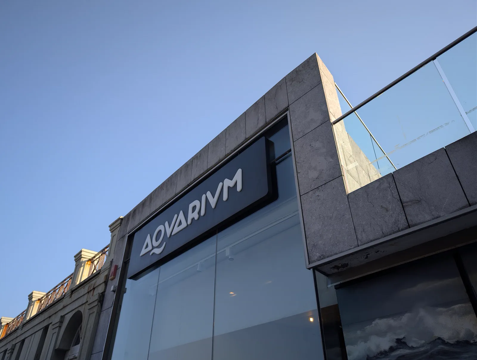 Fachada del Aquarium de San Sebastián con su rótulo «AQVARIVM» sobre el cristal, vista desde la calle al atardecer