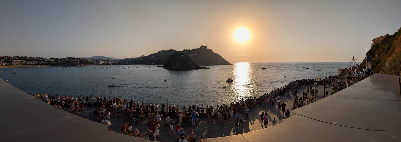 Panorámica de la bahía de la Concha desde la terraza del Aquarium, con la isla de Santa Clara en el centro, decenas de barcos fondeados y el Paseo Nuevo lleno de gente en primer plano