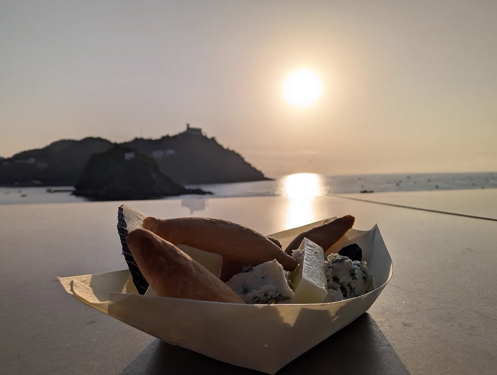 Barquilla de papel con tacos de queso, queso azul, picos y crackers apoyada en el muro de la terraza del Aquarium, con el monte Igueldo y el sol reflejado en el mar al fondo