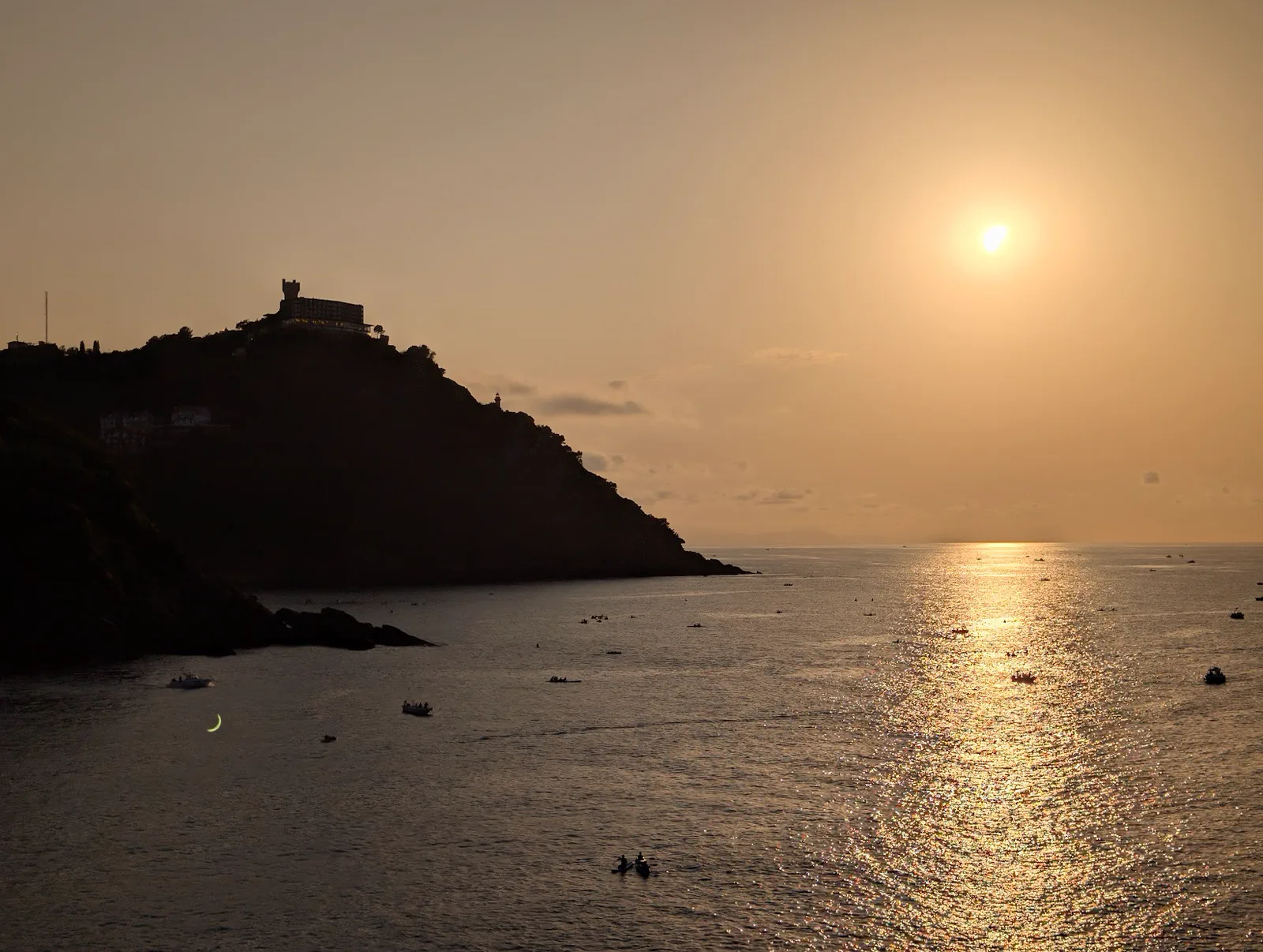 El monte Igueldo recortado a contraluz y el sol muy bajo sobre el mar, con decenas de embarcaciones esperando, cinco minutos antes del máximo del eclipse