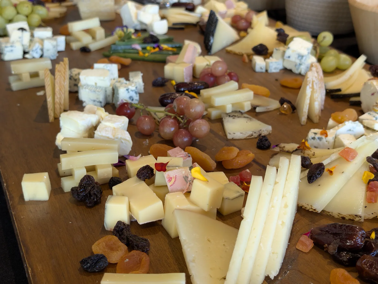 Gran tabla de quesos servida sobre madera en la terraza del Aquarium, con cuñas y tacos de distintos quesos, uvas, orejones, pasas y flores comestibles