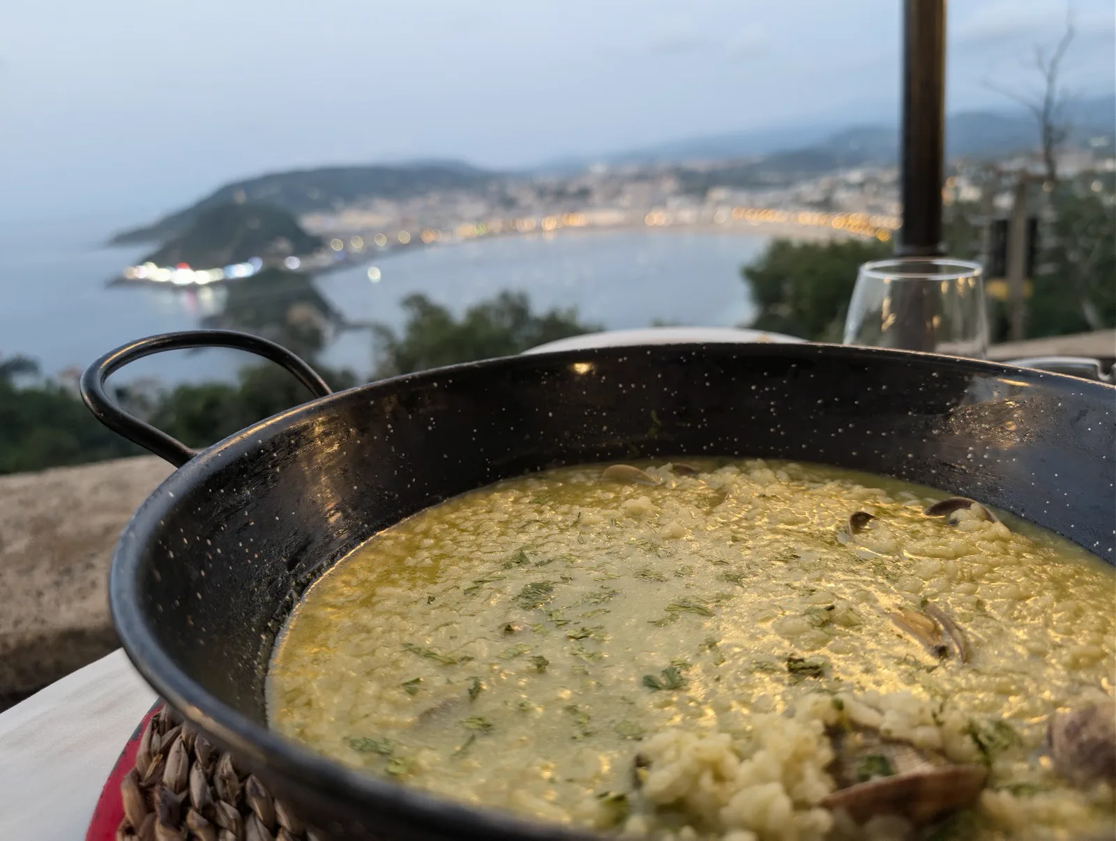Cazuela de arroz con almejas y perejil recién servida en la mesa del Asador Alaia, con la bahía de la Concha encendiéndose al anochecer detrás