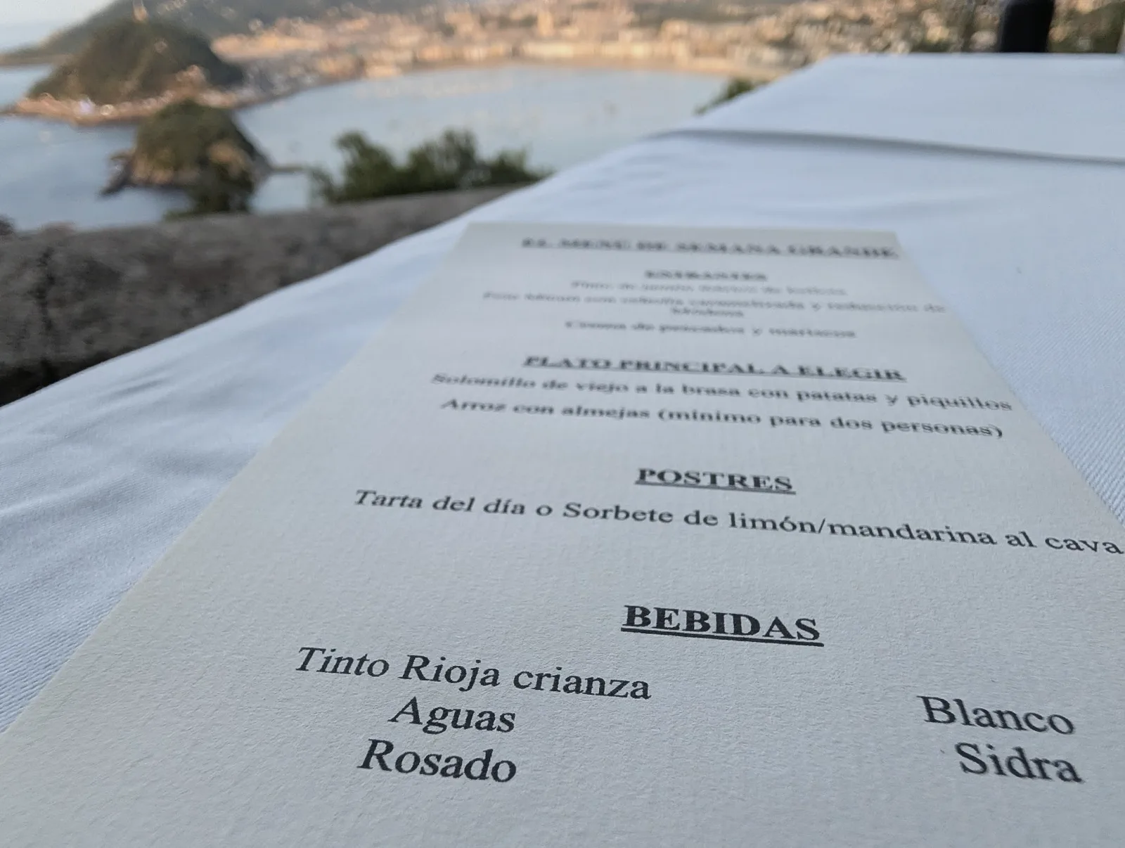La carta del menú de Semana Grande del Asador Alaia sobre el mantel, con los entrantes, el plato principal a elegir, los postres y las bebidas, y la bahía desenfocada al fondo