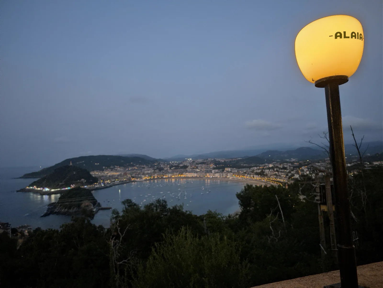 Farola encendida con el rótulo Alaia junto a la bahía de la Concha en hora azul, con las luces de la ciudad reflejadas en el agua