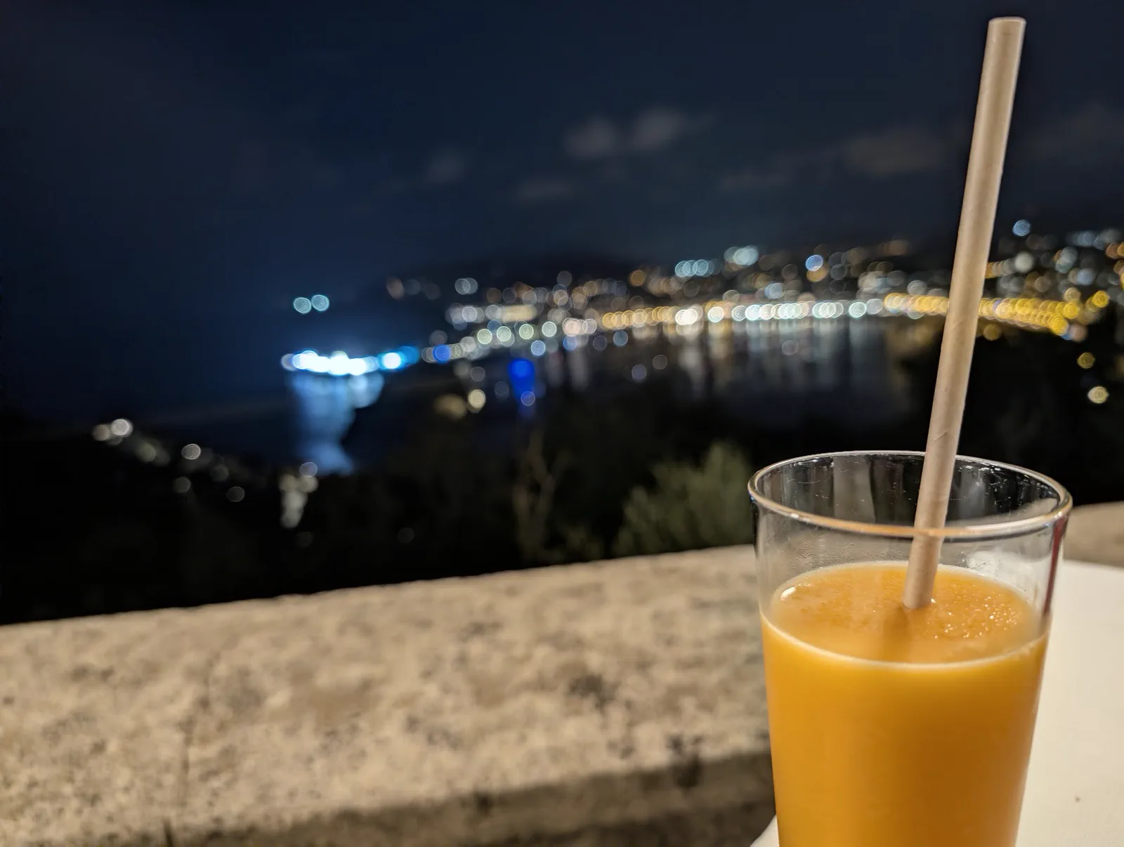Vaso de sorbete de mandarina con pajita apoyado en el muro de la terraza del Asador Alaia, con las luces de la bahía desenfocadas al fondo