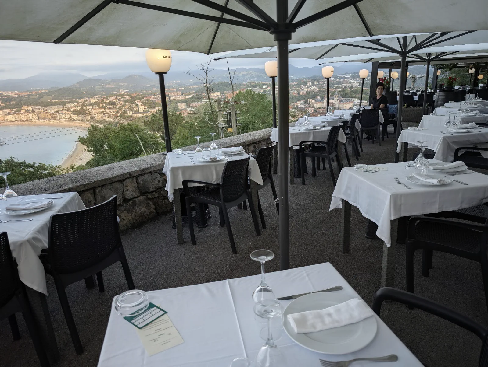 Terraza del Asador Alaia con las mesas puestas con mantel blanco bajo las sombrillas, y la playa de Ondarreta y la bahía de la Concha al fondo, más abajo