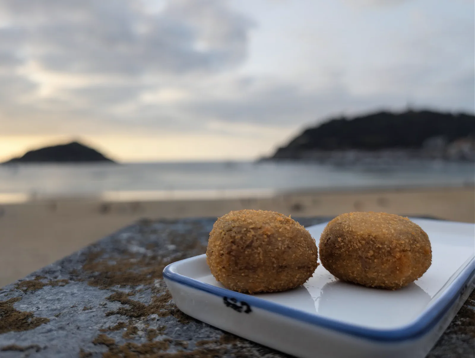 Dos croquetas en un plato rectangular apoyado sobre la barandilla de piedra, con la playa de la Concha y la isla de Santa Clara desenfocadas al fondo