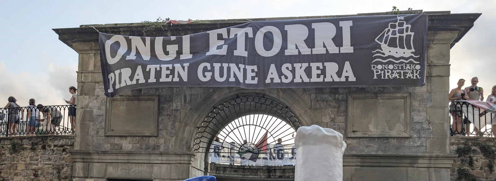Arco de piedra de la entrada al puerto de Donostia con una pancarta negra que dice «Ongi etorri piraten gune askera» y el barco pirata del logotipo de Donostiako Piratak