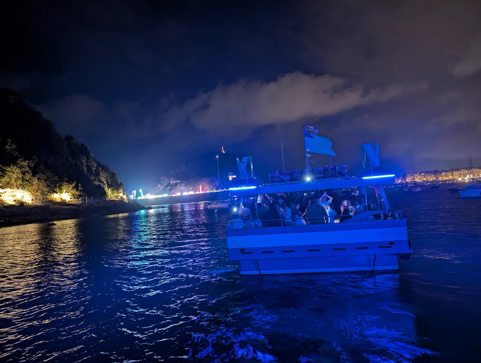 Una de las motoras navegando de noche por la bahía con los pasajeros a bordo bajo la tira de LED azul, con la costa oscura y las luces de la ciudad al fondo