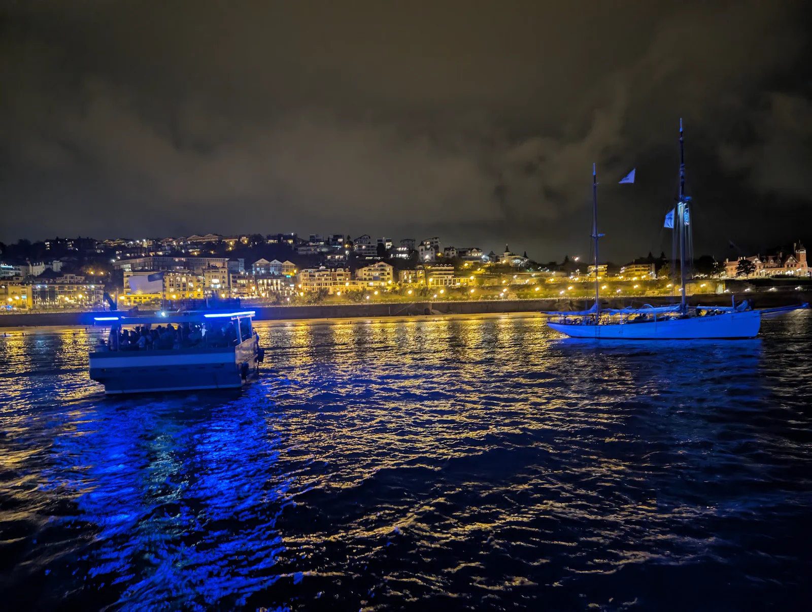 Una de las motoras navegando de noche por la bahía con su LED azul encendido, junto a un velero fondeado y con las luces de Ondarreta y la ladera de Igueldo al fondo
