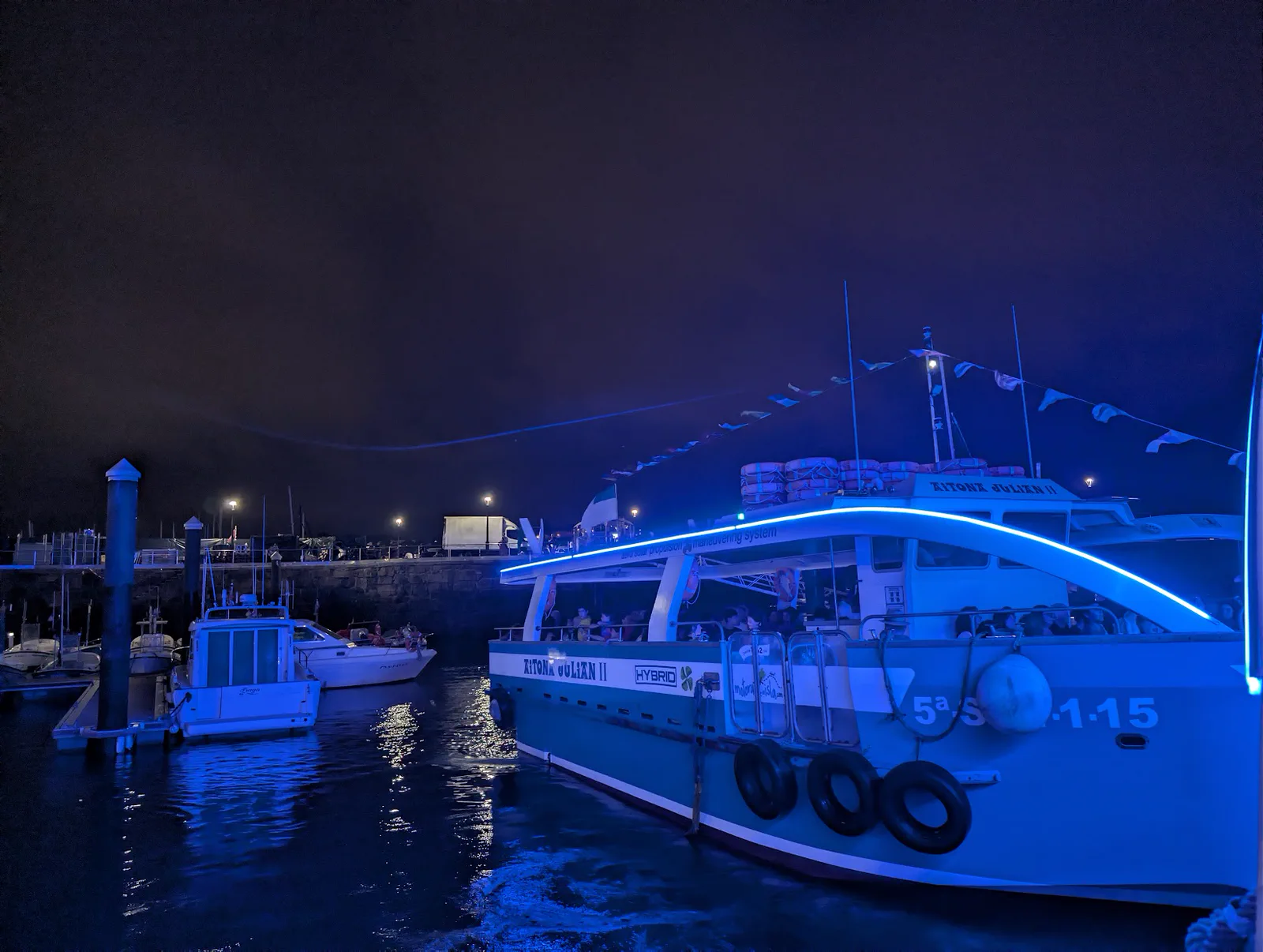 Una motora iluminada de azul saliendo del puerto de Donostia de noche, con los barcos de recreo amarrados y el muelle al fondo