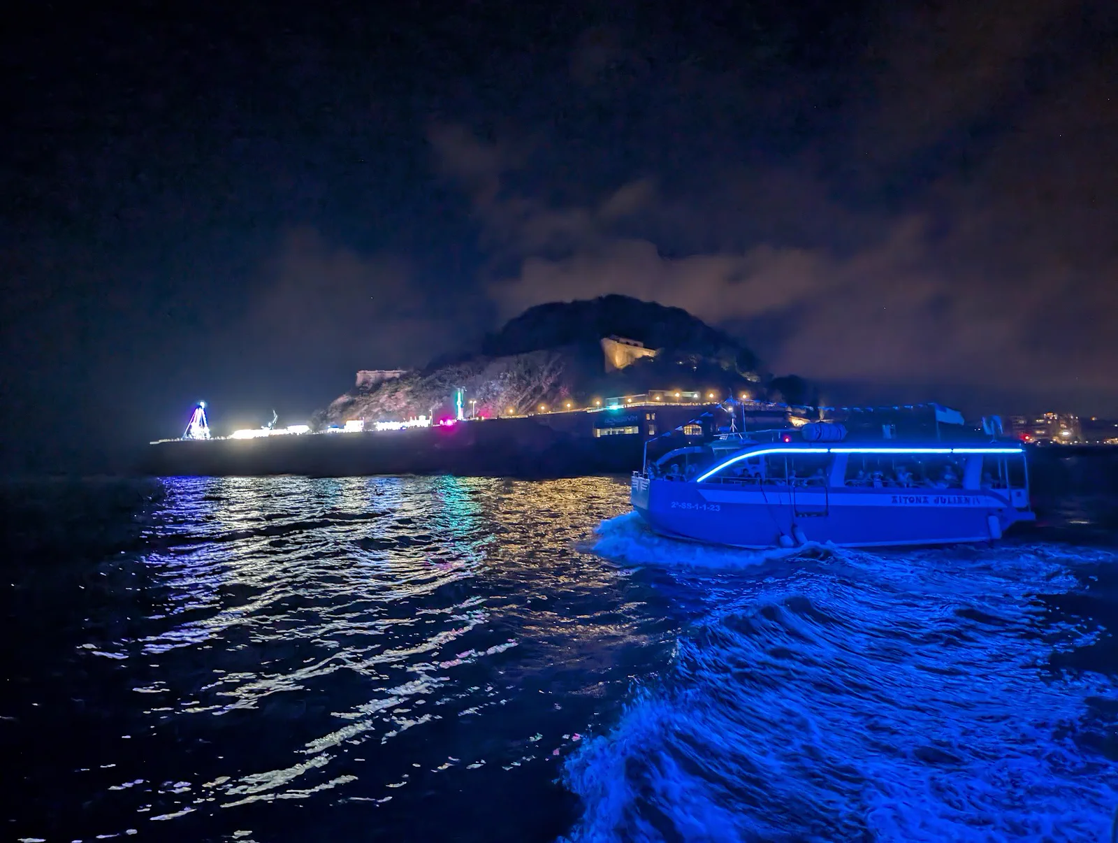 Otra de las motoras navegando de noche por la bahía con su tira de LED azul encendida, con el monte Urgull y las luces de las ferias del Paseo Nuevo al fondo